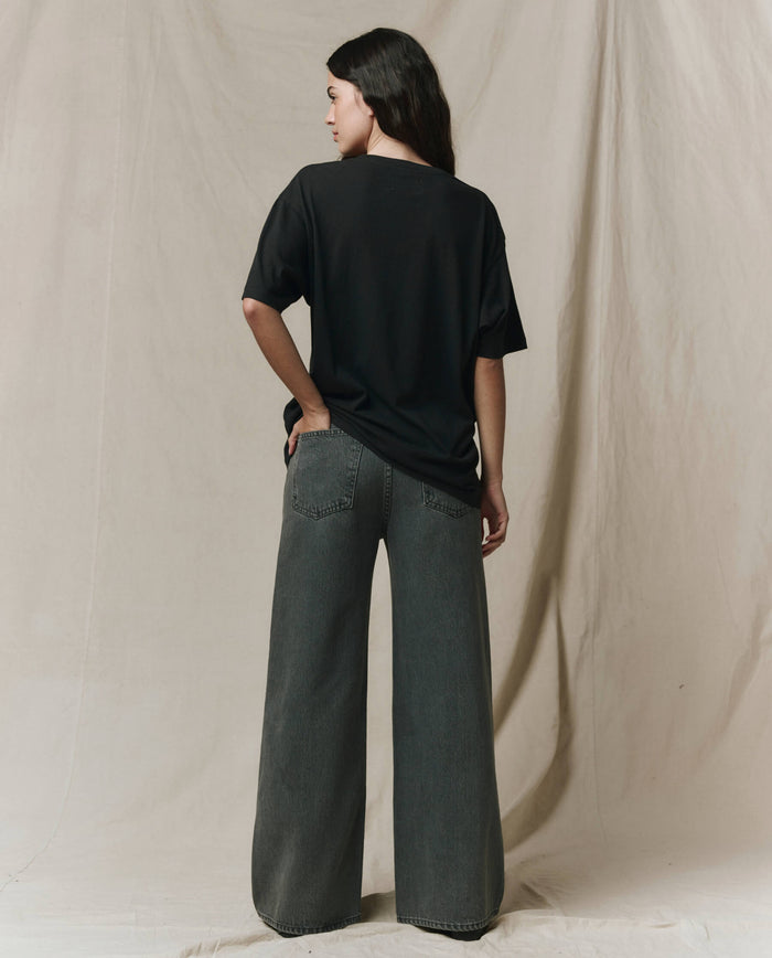A woman faces away, wearing THE GREAT. SP26 SEASONAL CORE The Slouchy Crew Tee in Almost Black—an oversized fit—paired with wide-leg gray jeans, set against a neutral fabric backdrop.