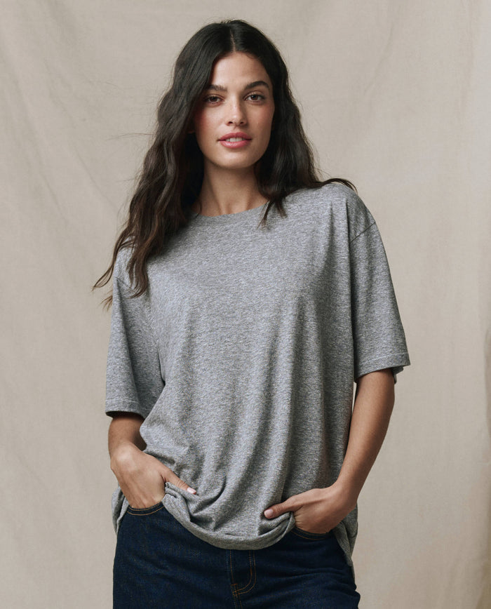 A woman with long dark hair wears THE GREAT.'s The Slouchy Crew Tee in Heather Grey (SP26 SEASONAL CORE), paired with dark jeans, standing with her hands in her pockets against a plain beige background.