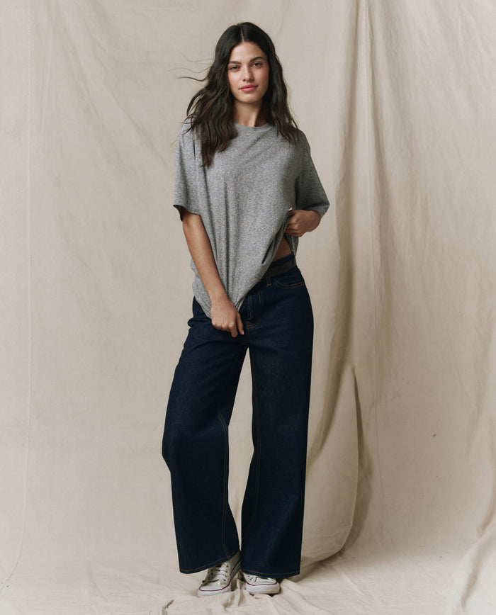 A person stands against a beige backdrop, wearing THE GREAT. SP26 SEASONAL CORE The Slouchy Crew Tee in Heather Grey, paired with dark wide-leg jeans and white sneakers.