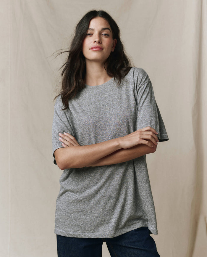 A person stands with arms crossed, wearing THE GREAT. SP26 SEASONAL CORE The Slouchy Crew Tee in Heather Grey and dark pants, against a neutral fabric backdrop.