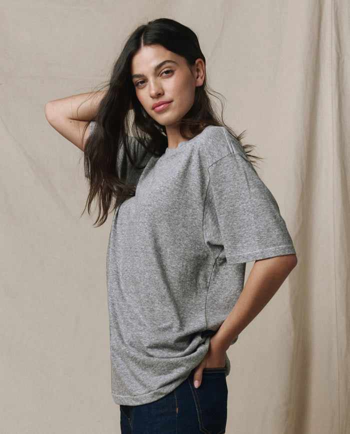 A woman with long dark hair models THE GREAT.'s The Slouchy Crew Tee in Heather Grey, styled oversized with blue jeans. She stands before a beige backdrop, hand behind her head and the other in her pocket.