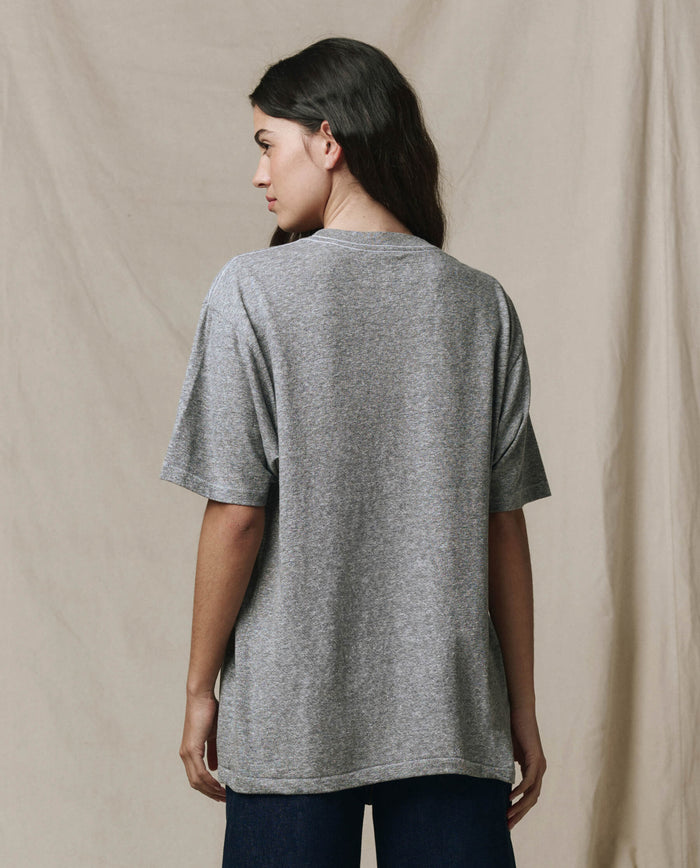 A person with long dark hair, wearing THE GREAT. SP26 SEASONAL CORE The Slouchy Crew Tee in Heather Grey, and dark pants stands facing away from the camera against a beige fabric backdrop.