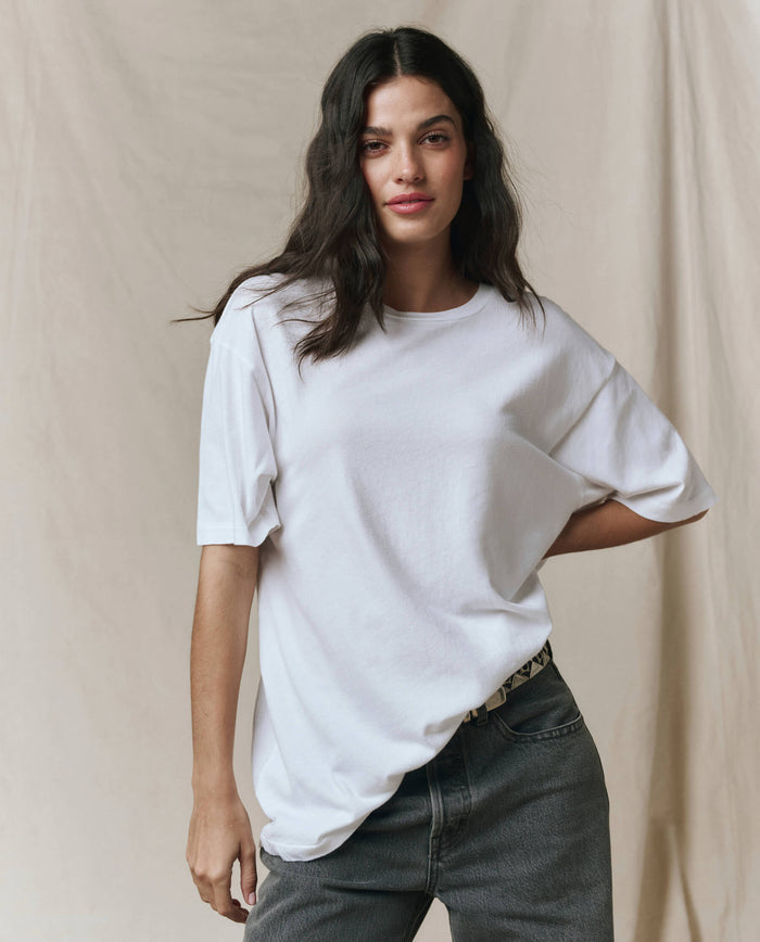 A person with long dark hair wears THE GREAT. SP26 SEASONAL CORE The Slouchy Crew Tee in True White, styled oversized with dark jeans, standing before a neutral beige backdrop.
