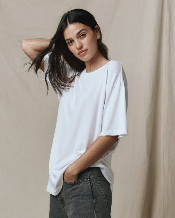 A person with long dark hair wears THE GREAT. SP26 SEASONAL CORE The Slouchy Crew Tee in True White, styled oversized with jeans, standing before a beige backdrop, one hand behind their head and the other in a pocket.