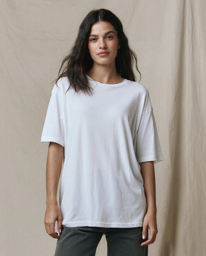 A person with long dark hair wears THE GREAT.’s The Slouchy Crew Tee in True White (SP26 SEASONAL CORE) and dark pants, standing before a beige fabric backdrop.