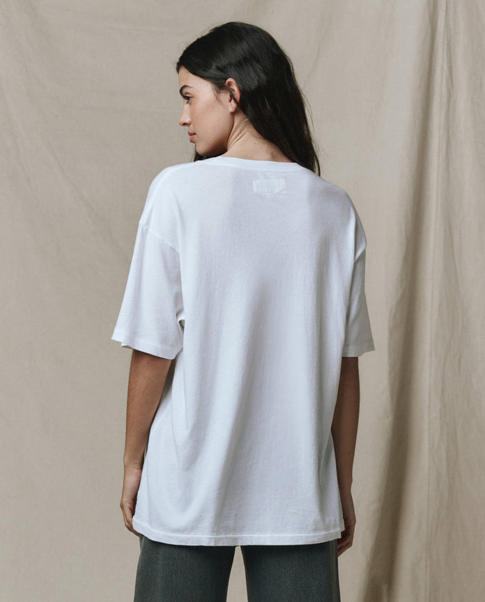 A person with long dark hair stands with their back to the camera, wearing THE GREAT.’s The Slouchy Crew Tee. in True White (SP26 SEASONAL CORE), styled oversized with dark pants, against a beige fabric backdrop.