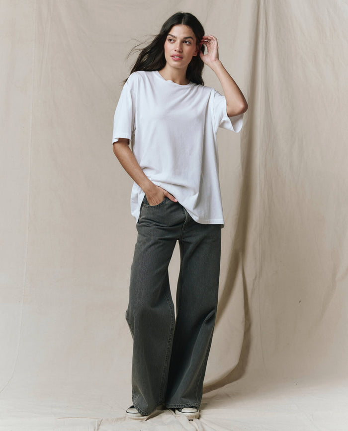 A woman stands against a beige fabric backdrop, wearing THE GREAT. SP26 SEASONAL CORE The Slouchy Crew Tee in True White, styled with wide-leg dark jeans and sneakers. One hand is in her pocket, the other touches her hair.