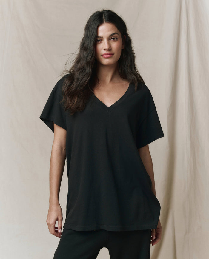 A woman with long dark hair wears THE GREAT.'s The Slouchy V-Neck Tee in Almost Black from the SP26 SEASONAL CORE, paired with black pants, standing against a plain beige backdrop.