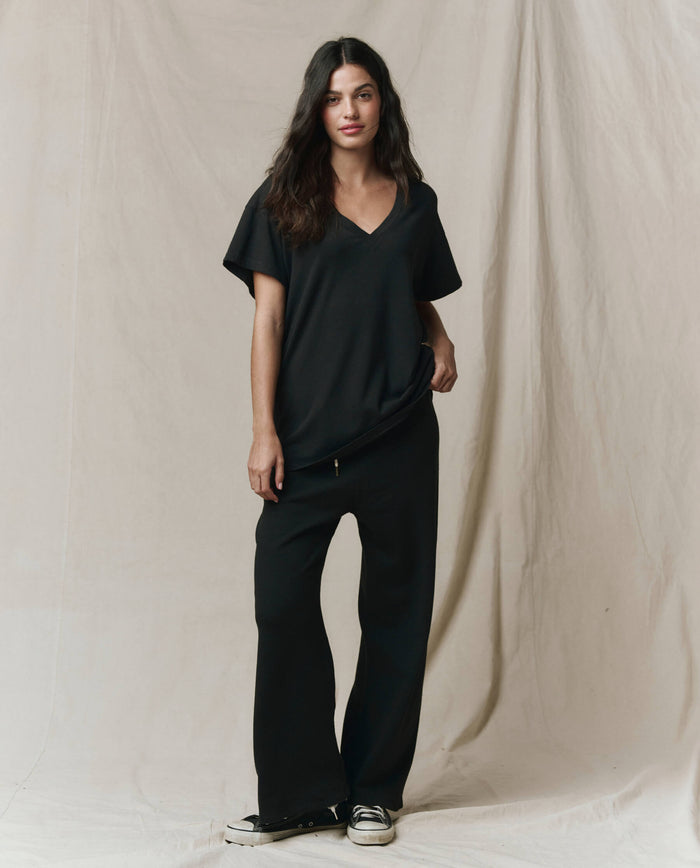 A person stands on a beige fabric backdrop wearing THE GREAT. SP26 SEASONAL CORE The Slouchy V-Neck Tee in Almost Black, styled with wide-leg black pants and black-and-white sneakers.
