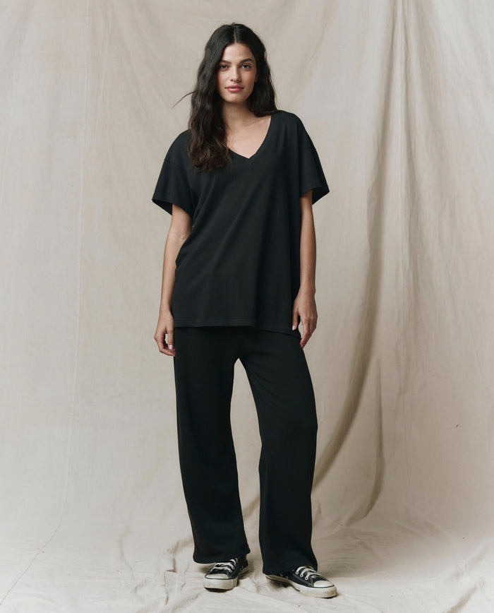 A woman stands before a beige backdrop, wearing THE GREAT.'s The Slouchy V-Neck Tee in Almost Black from the SP26 SEASONAL CORE, paired with black wide-leg pants and black sneakers with white soles.