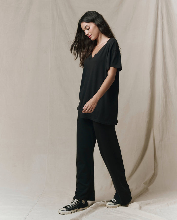 A woman stands against a beige backdrop, wearing THE GREAT. SP26 SEASONAL CORE The Slouchy V-Neck Tee in Almost Black, paired with black pants and sneakers.