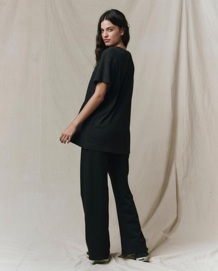 A person stands sideways, looking over their shoulder, wearing THE GREAT.'s The Slouchy V-Neck Tee in Almost Black from the SP26 SEASONAL CORE collection and black wide-leg pants against a neutral draped fabric background.