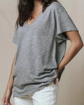 A person wears THE GREAT.’s The Slouchy V-Neck Tee in Heather Grey from the SP26 SEASONAL CORE, styled with white pants and one hand in their pocket, standing against a plain background.