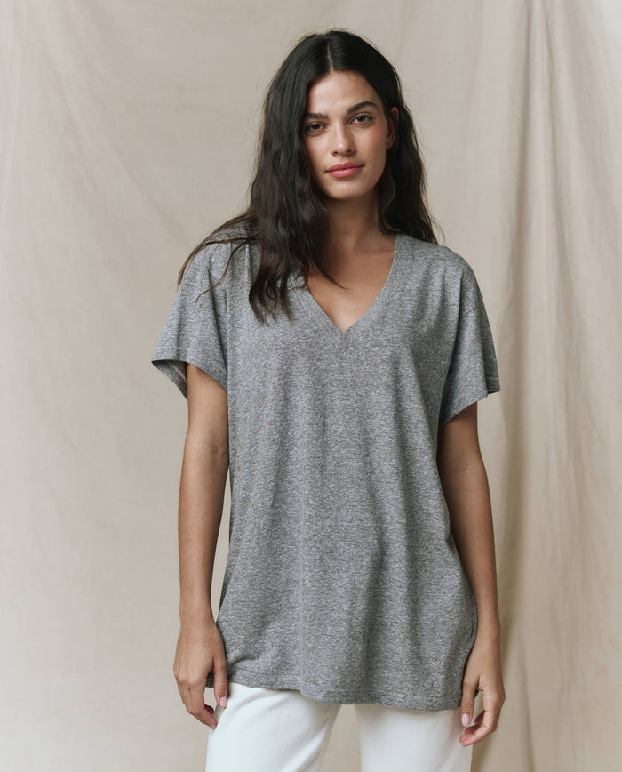 A woman with long dark hair wears THE GREAT. SP26 SEASONAL CORE's The Slouchy V-Neck Tee in Heather Grey and white pants, standing before a beige fabric backdrop.