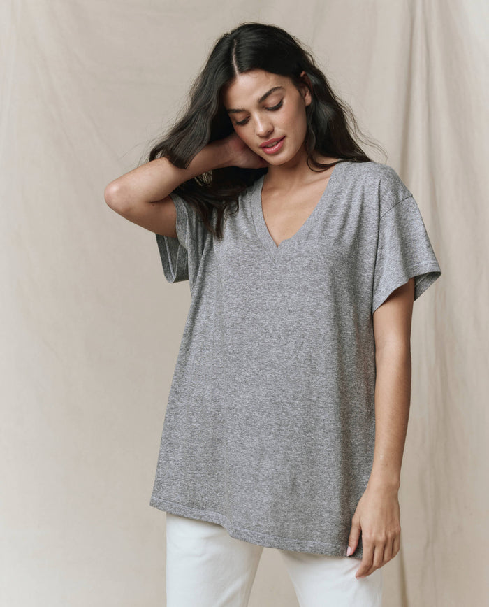 A woman wears THE GREAT. SP26 SEASONAL CORE The Slouchy V-Neck Tee in Heather Grey with white pants, standing against a neutral beige background.