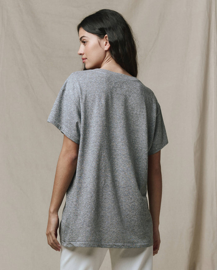 A woman faces away, wearing THE GREAT.'s The Slouchy V-Neck Tee in Heather Grey—an oversized, short-sleeved washed jersey top—from the SP26 SEASONAL CORE collection, paired with white pants, set against a neutral fabric backdrop.