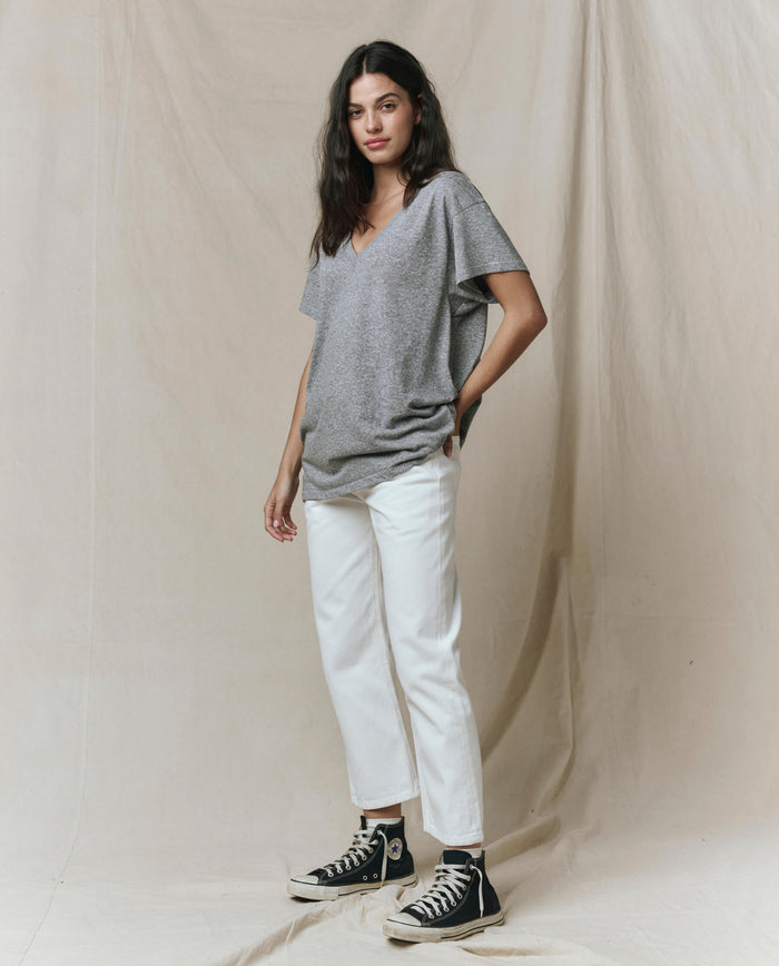 A woman stands before a beige backdrop, wearing THE GREAT. SP26 SEASONAL CORE’s The Slouchy V-Neck Tee in heather grey, paired with white pants and black high-top sneakers.