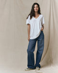 A person stands against a beige fabric backdrop, wearing THE GREAT. SP26 SEASONAL CORE The Slouchy V-Neck Tee in True White, paired with wide-leg blue jeans and light-colored sneakers.