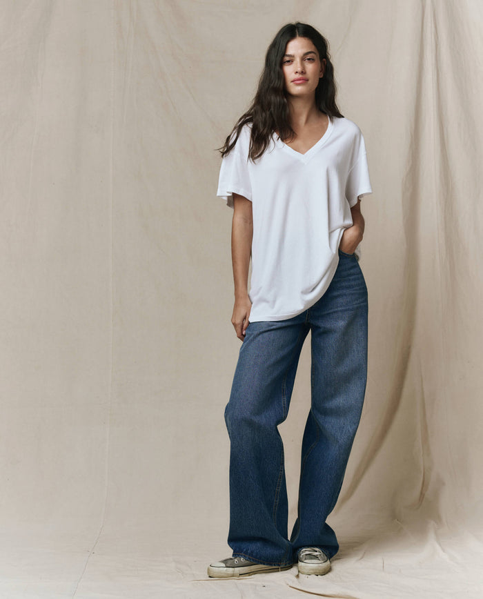 A person stands against a beige fabric backdrop, wearing THE GREAT. SP26 SEASONAL CORE The Slouchy V-Neck Tee in True White, paired with wide-leg blue jeans and light-colored sneakers.