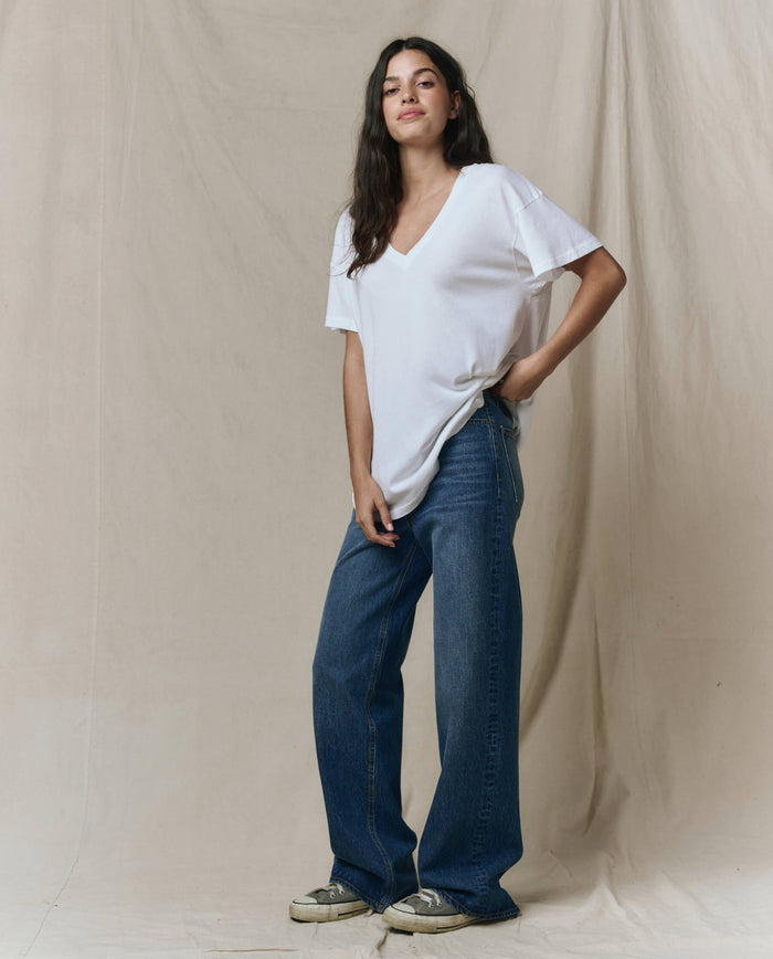 A person stands against a beige fabric backdrop, wearing THE GREAT. SP26 SEASONAL CORE The Slouchy V-Neck Tee in True White, wide-leg blue jeans, and sneakers.