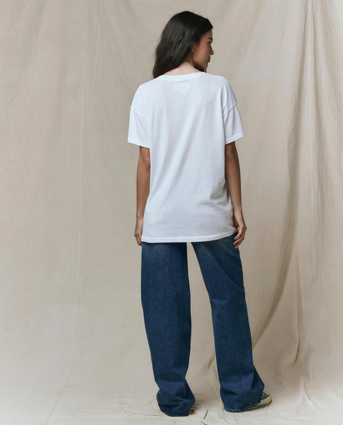 A person faces away, wearing THE GREAT. SP26 SEASONAL CORE's The Slouchy V-Neck Tee in True White—an oversized washed jersey—paired with loose blue jeans, set against a neutral fabric backdrop.