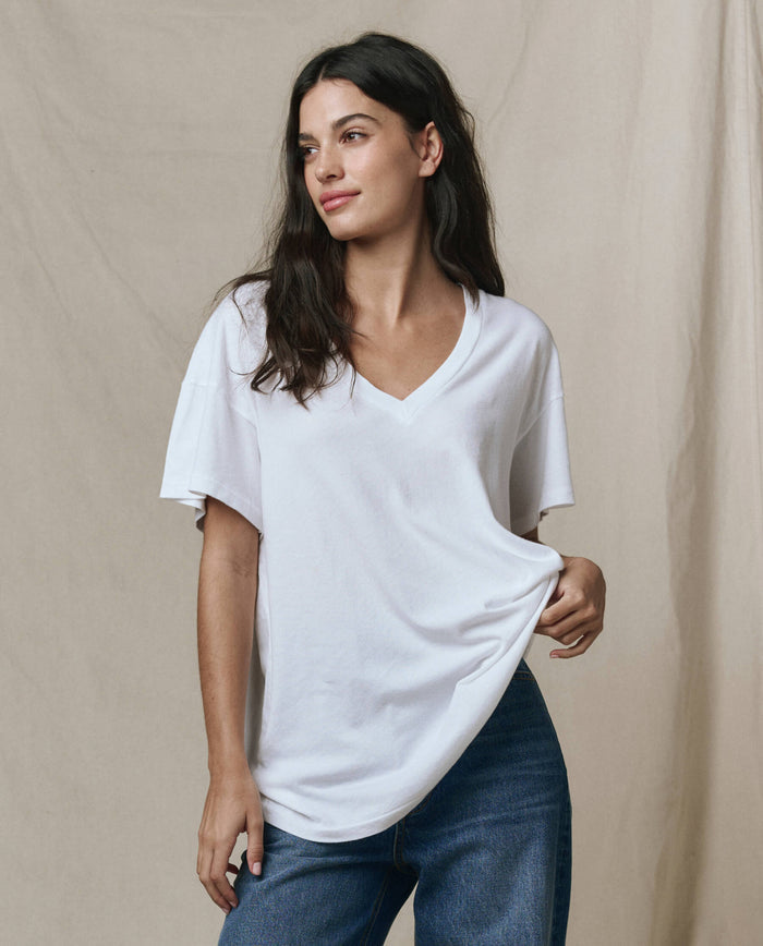 A woman wears THE GREAT.'s The Slouchy V-Neck Tee in True White from the SP26 SEASONAL CORE, paired with blue jeans, standing before a beige backdrop and holding her shirt’s hem.