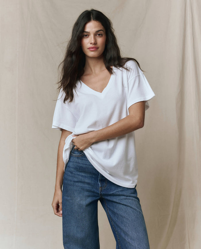 A woman in THE GREAT. SP26 SEASONAL CORE The Slouchy V-Neck Tee in True White and blue jeans stands before a beige fabric backdrop, looking at the camera.