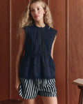 A woman with long blonde hair wears THE GREAT.'s The Sonnet Top in navy and black-and-white striped shorts, standing in front of wooden paneling.