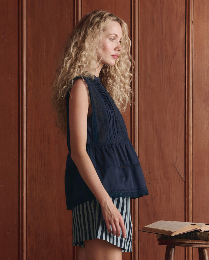A woman with long curly blonde hair stands in profile, wearing THE GREAT. SP26 D1 The Sonnet Top in Navy and striped shorts beside a small table with an open book, set against wood paneling.