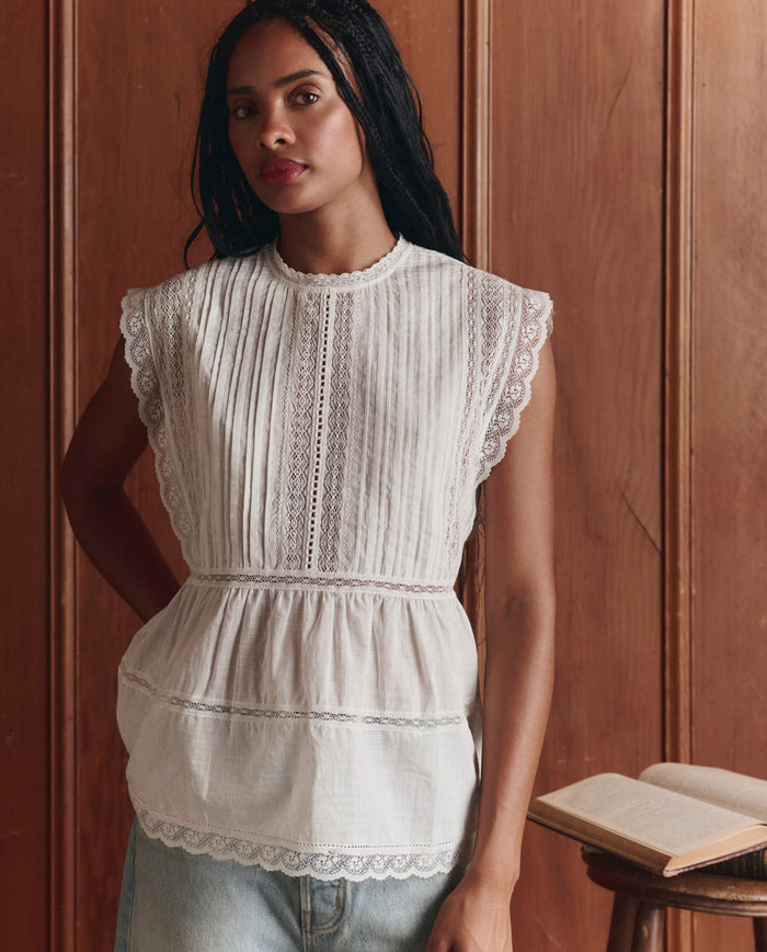 A woman with long braided hair wears THE GREAT.'s Sonnet Top in True White, paired with light blue jeans, standing by a wooden wall with an open book on a table beside her.