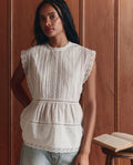 A woman with long braided hair wears THE GREAT.'s Sonnet Top in True White, paired with light blue jeans, standing by a wooden wall with an open book on a table beside her.