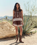 A woman stands outdoors among rocks, agave plants, and mountains, wearing THE GREAT. PF26's The Sparrow Short in Chocolate with cowboy boots and a sleeveless patterned top.