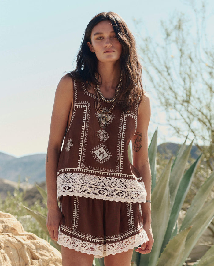 A woman poses outdoors in THE GREAT. PF26 The Sparrow Short—Chocolate, paired with a brown sleeveless top and layered necklaces. White lace trim accents the shorts; desert plants and hills form the backdrop.