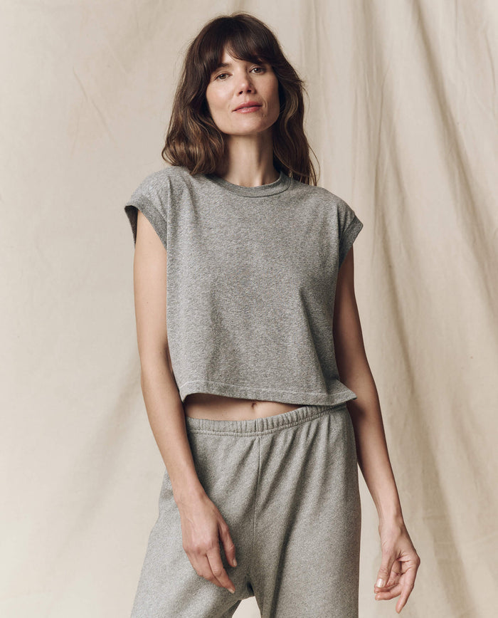 A woman stands against a plain fabric backdrop, featuring "The Square Tee" in heather grey by THE GREAT. SU23 CORE KNITS, paired with matching sweatpants.