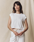 A person with long hair stands against a plain background, confidently wearing THE GREAT. SU23 CORE KNITS’ The Square Tee in True White along with white pants, looking at the camera.