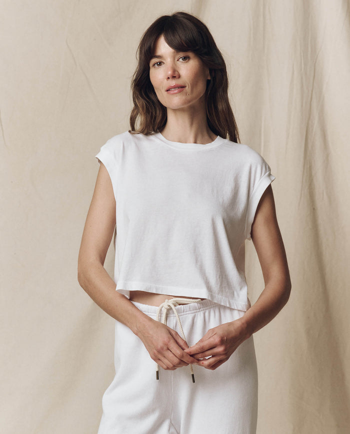 A person with long hair stands against a plain background, confidently wearing THE GREAT. SU23 CORE KNITS’ The Square Tee in True White along with white pants, looking at the camera.