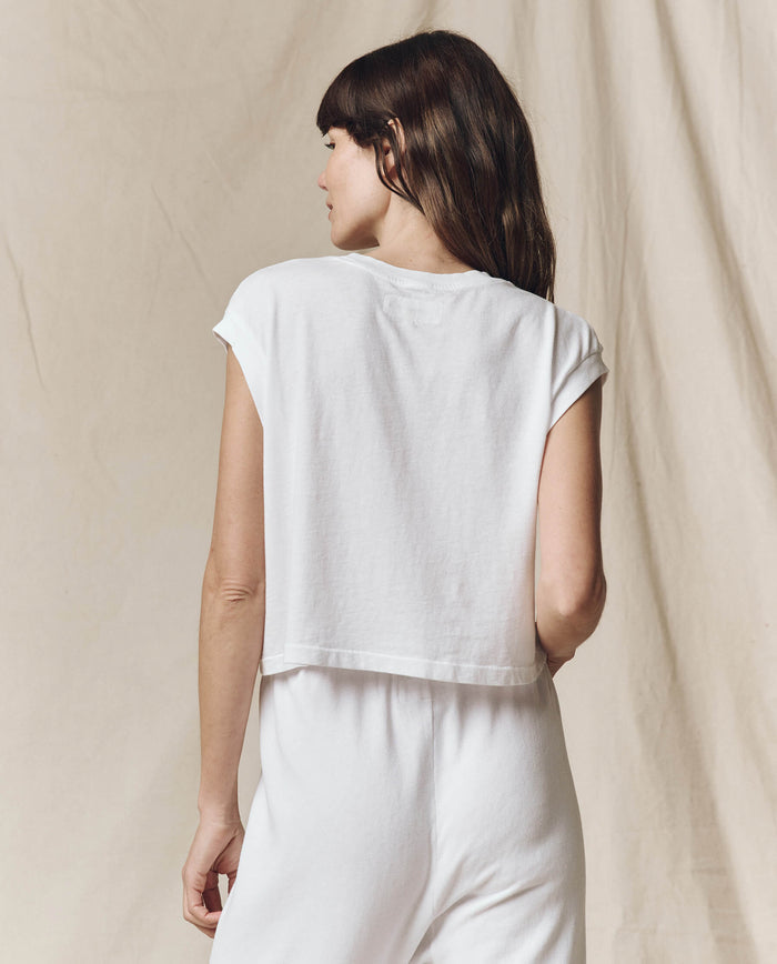 With long, dark hair turned away, a person stands against a beige backdrop wearing THE GREAT. SU23 CORE KNITS' The Square Tee in True White and high-waisted denim.