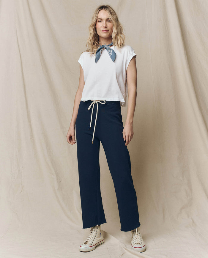Against a beige backdrop, a person wears The Square Tee in True White by THE GREAT. SU23 CORE KNITS, paired with a blue scarf, dark pants, and white sneakers.