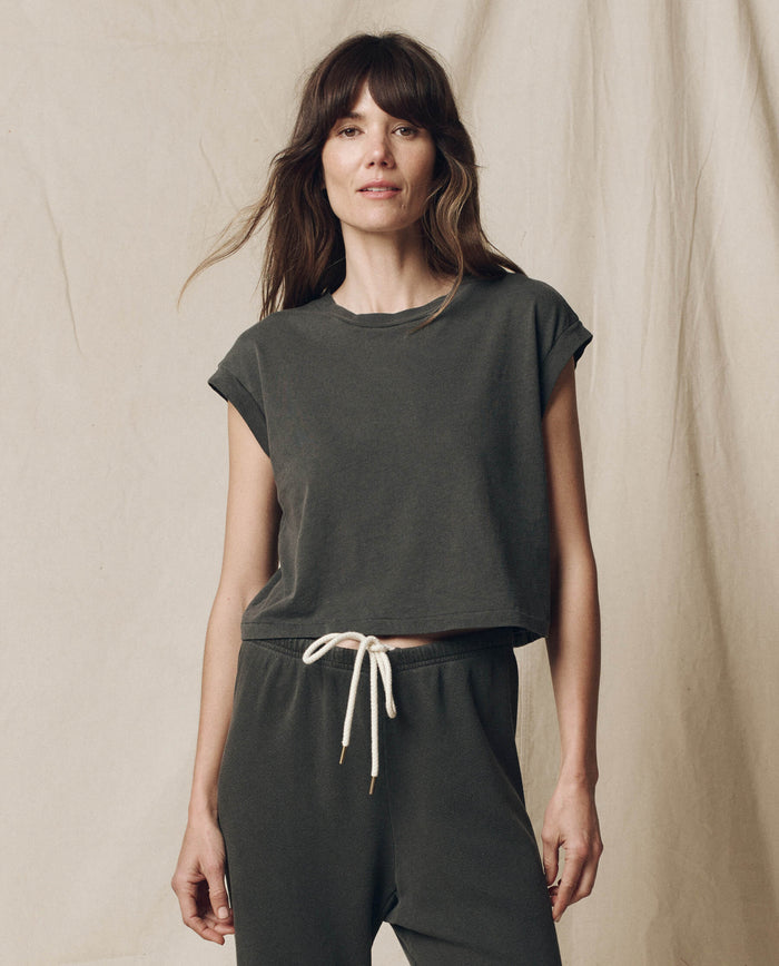 A woman with long brown hair stands against a plain backdrop, wearing The Square Tee in Washed Black by THE GREAT. SU23 CORE KNITS and matching drawstring pants.