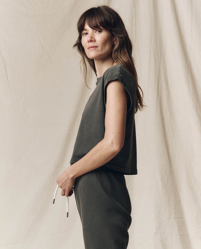 A person with long brown hair stands sideways, wearing The Square Tee in Washed Black by THE GREAT. SU23 CORE KNITS and high-waisted denim, holding eyeglasses against a neutral fabric backdrop.