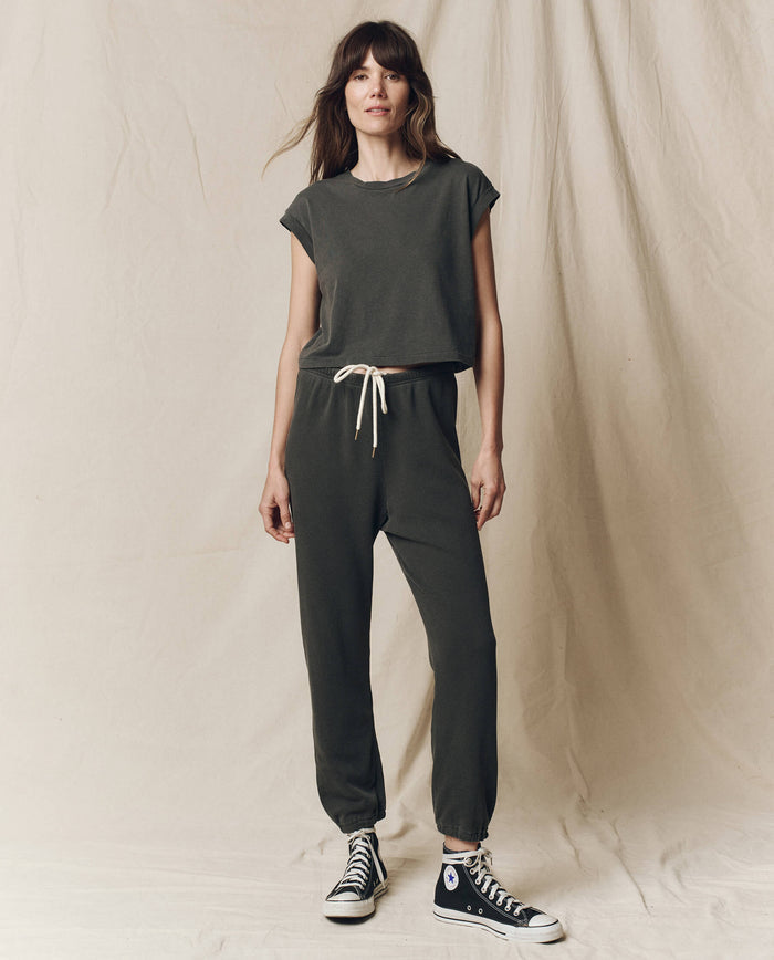 Someone in a casual dark outfit featuring a vintage athletic tee, likely "The Square Tee" in Washed Black by THE GREAT. SU23 CORE KNITS, joggers, and black high-top sneakers, stands against a beige fabric backdrop.