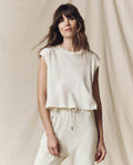 A person with long hair wears THE GREAT.'s handcrafted The Square Tee in Washed White, standing in front of a light-colored fabric backdrop.