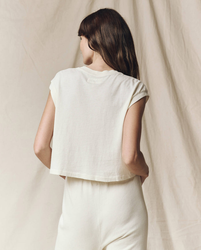 A person with long hair stands against a beige backdrop, wearing The Square Tee in Washed White with matching pants, crafted by THE GREAT. SU23 CORE KNITS in L.A.