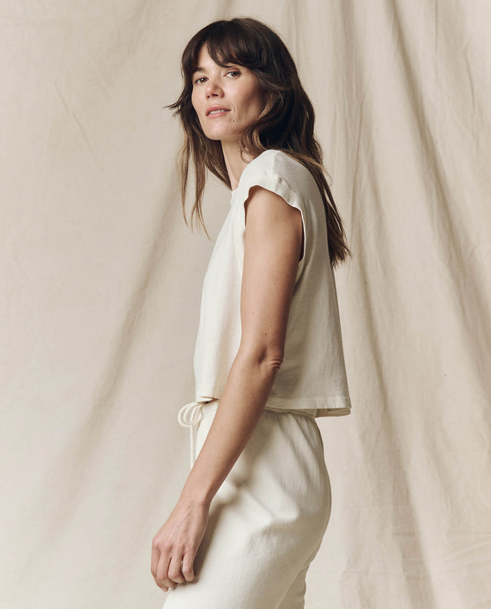 In front of a neutral fabric backdrop, a long-haired individual in THE GREAT. SU23 CORE KNITS' Washed White Square Tee and pants gazes to the side.