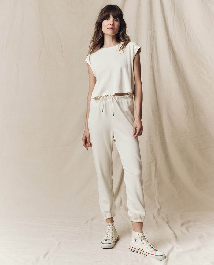 Wearing 'The Square Tee' in Washed White by THE GREAT. SU23 CORE KNITS and ankle-length pants, a person stands against a beige fabric backdrop, highlighting their high-top sneakers. The outfit embodies handcrafted elegance reminiscent of L.A., blending modern style with timeless charm.