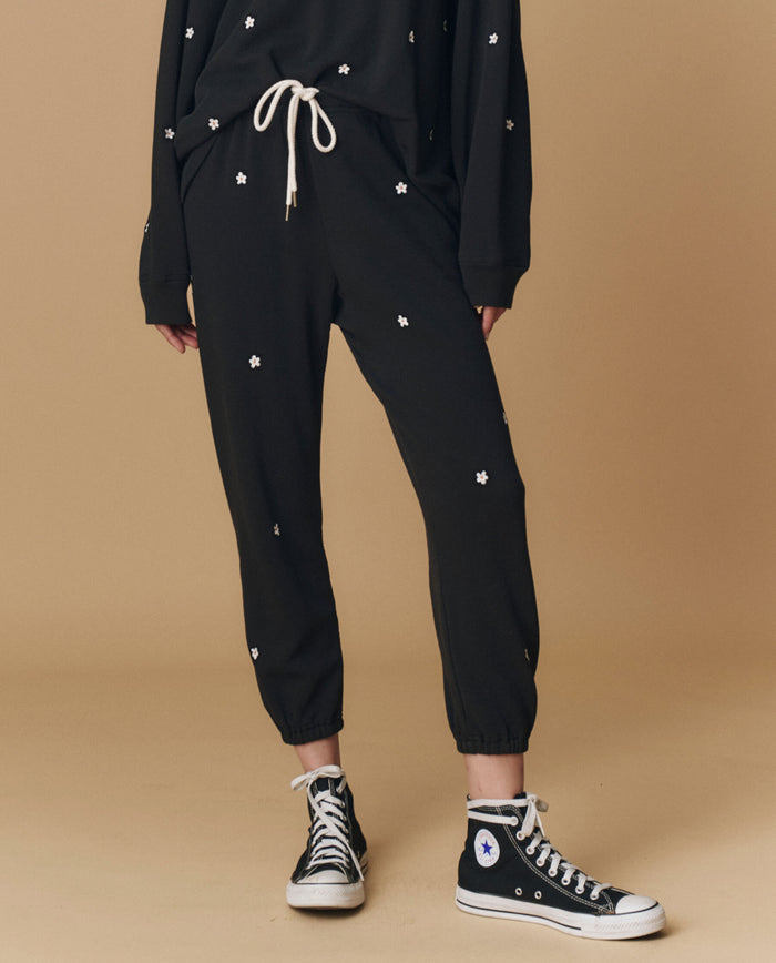 A person wears THE GREAT. PF25 EMB KNITS The Stadium Sweatpant. Embroidered -- Almost Black With Daisy Embroidery, a matching sweatshirt, and black high-top Converse sneakers, standing against a beige background.