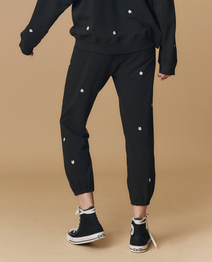 A person stands on a beige background wearing THE GREAT. PF25 EMB KNITS The Stadium Sweatpant. Embroidered in Almost Black With Daisy Embroidery, paired with a matching sweatshirt and black high-top sneakers.