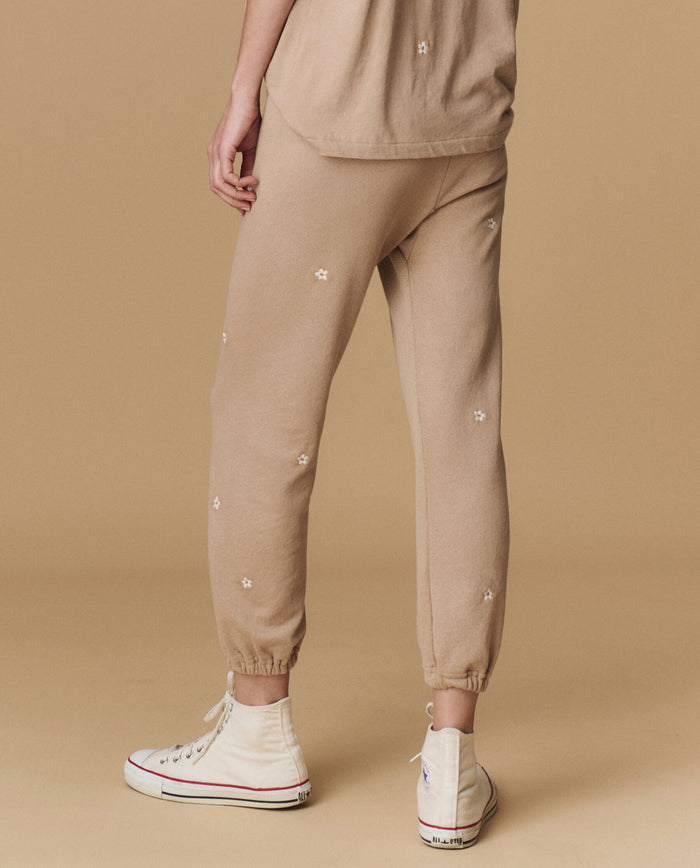 A person wears THE GREAT. PF25 EMB KNITS The Stadium Sweatpant, Embroidered—Khaki with Daisy Embroidery—with a matching tan shirt and white high-top sneakers, standing against a beige background.