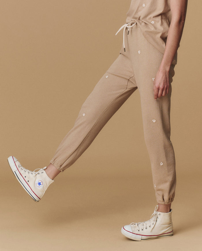 A person wears THE GREAT. PF25 EMB KNITS The Stadium Sweatpant. Embroidered in khaki with daisy embroidery, a matching top, and off-white high-top Converse sneakers, standing against a beige background.