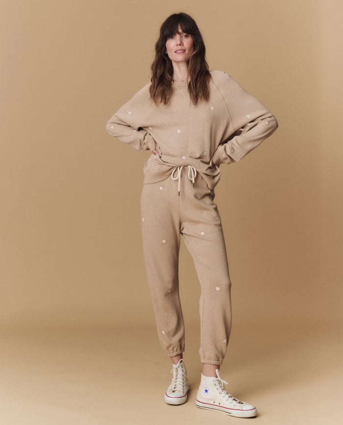 A woman wears THE GREAT. PF25 EMB KNITS’ The Stadium Sweatpant. Embroidered in khaki with daisy embroidery, paired with a matching sweatshirt and white high-top sneakers, standing against a beige backdrop.